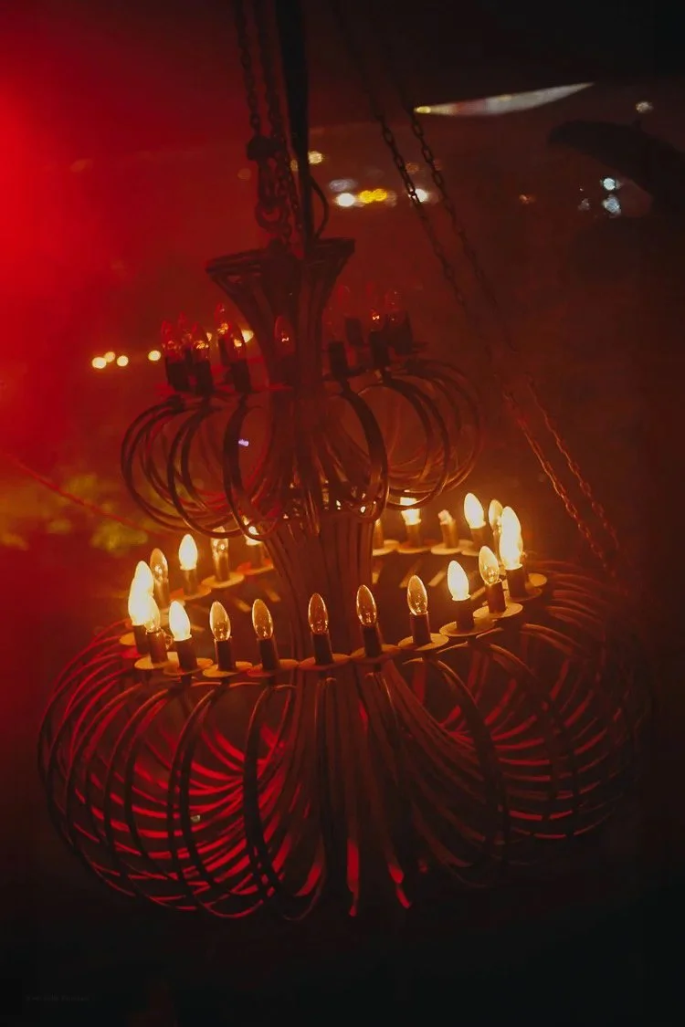 A chandelier with multiple lights, made of curved metal wires, hanging from the ceiling in a dimly lit room with red lighting.