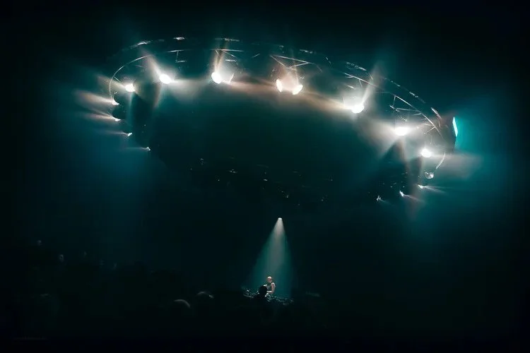 A DJ performing on a stage with a large, illuminated, circular stage lighting structure above him, in a dark nightclub or concert venue.
