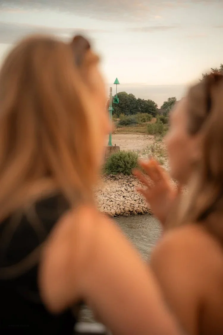 Two women near a body of water, with one gesturing as they look towards a green navigational marker on the water, with trees and an overcast sky in the background.