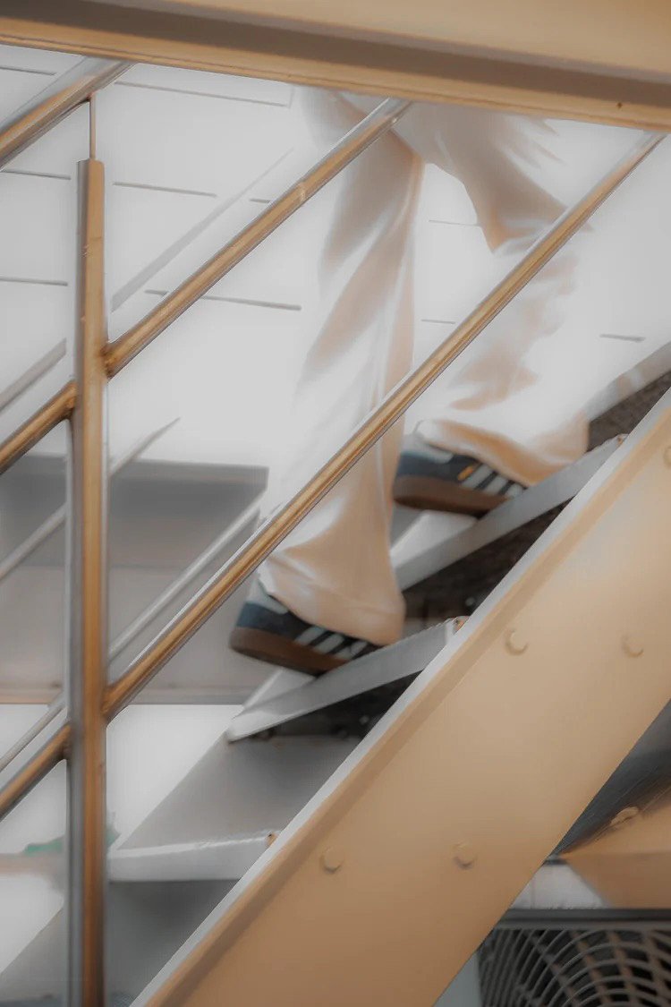 A person in beige pants and navy-blue sneakers with white stripes climbing metal stairs with wooden handrails, viewed from below.