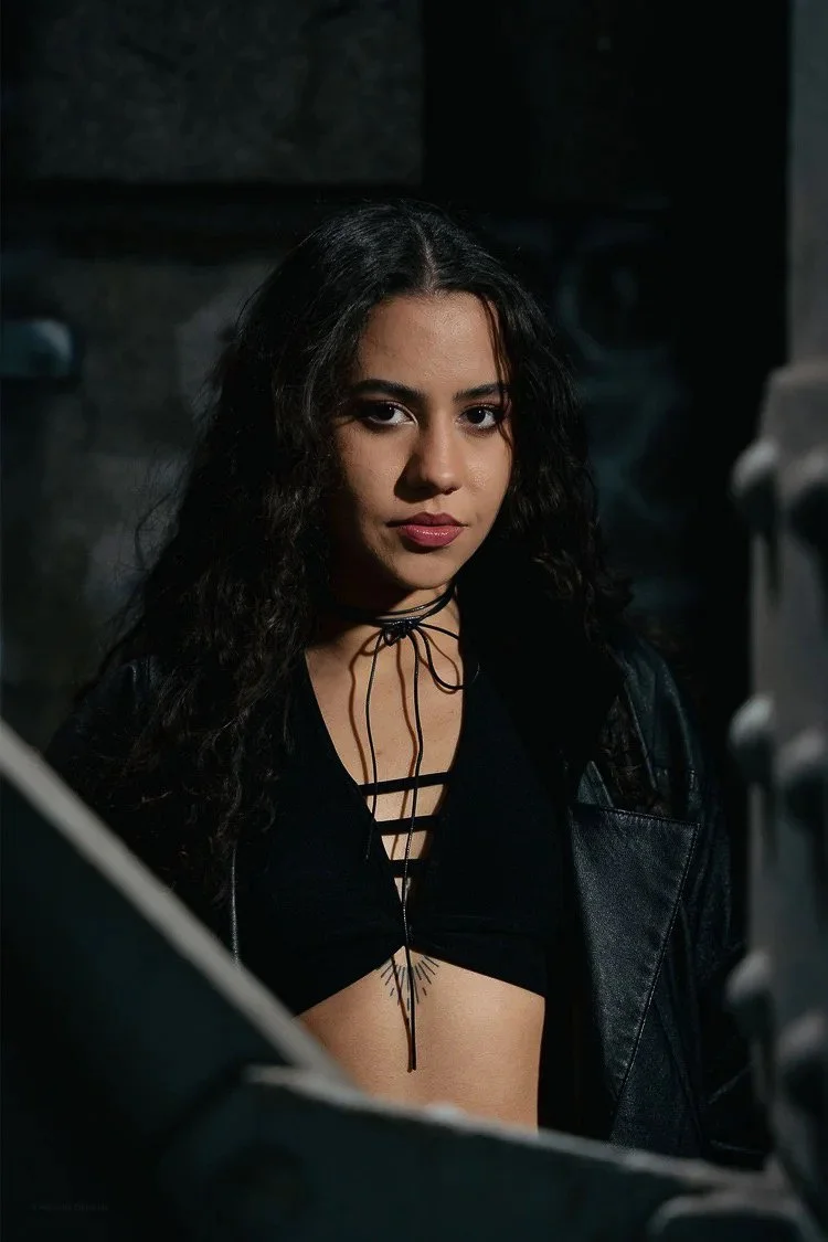 Young woman with wavy dark hair, wearing a black lace-up top and a black leather jacket, standing in a dark, industrial environment.