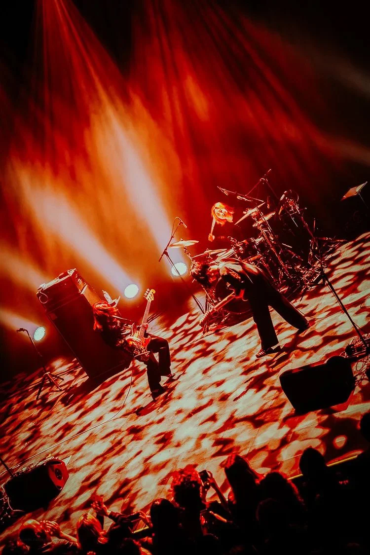 Musicians performing on stage with orange and red lighting, playing guitars and drums, audience watching.