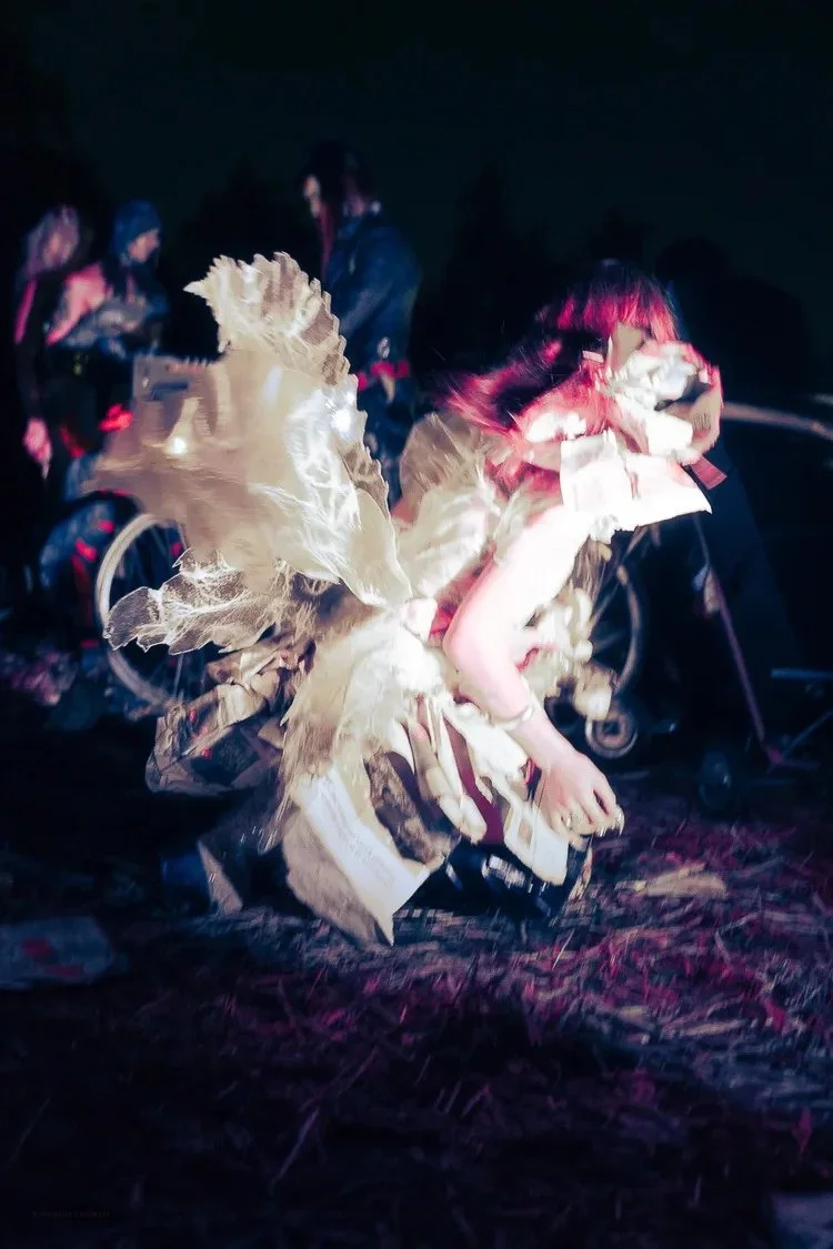 Person wearing an elaborate costume with large feathered wings and a white dress, crouching on the ground at night with other people and bikes in the background.