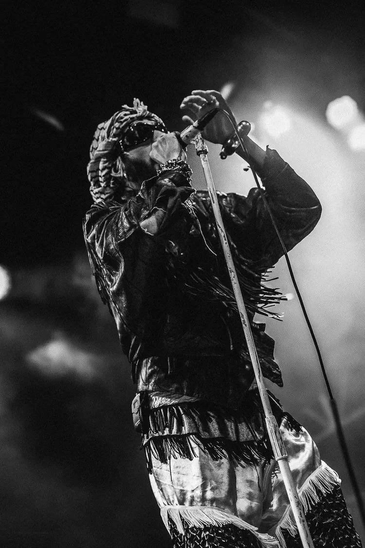 A black and white photograph of a singer on stage, wearing a fringed leather jacket, patterned pants, sunglasses, and a patterned headscarf, holding a microphone on a stand.