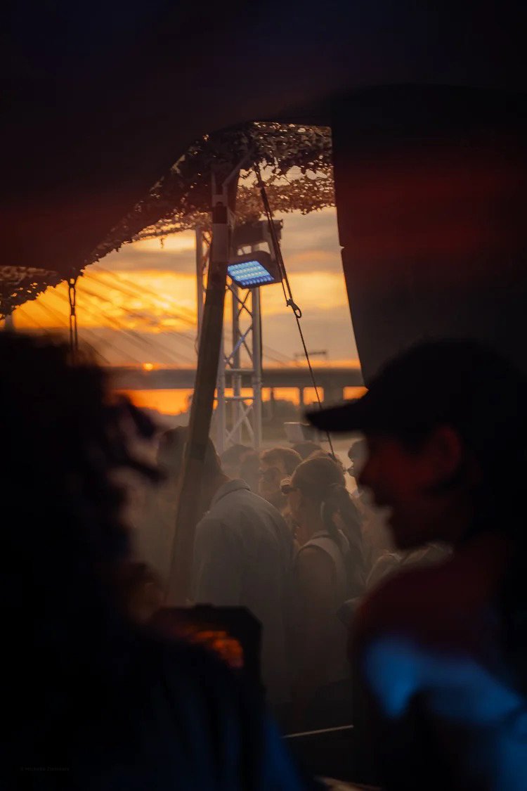 People gathered outdoors during sunset, seen through a window or opening, with a view of the sky and structures in the background.