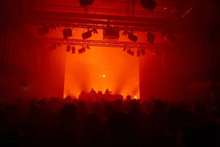 A concert or live music event with a large crowd, dimly lit with orange stage lighting and silhouettes of DJs or performers on stage.