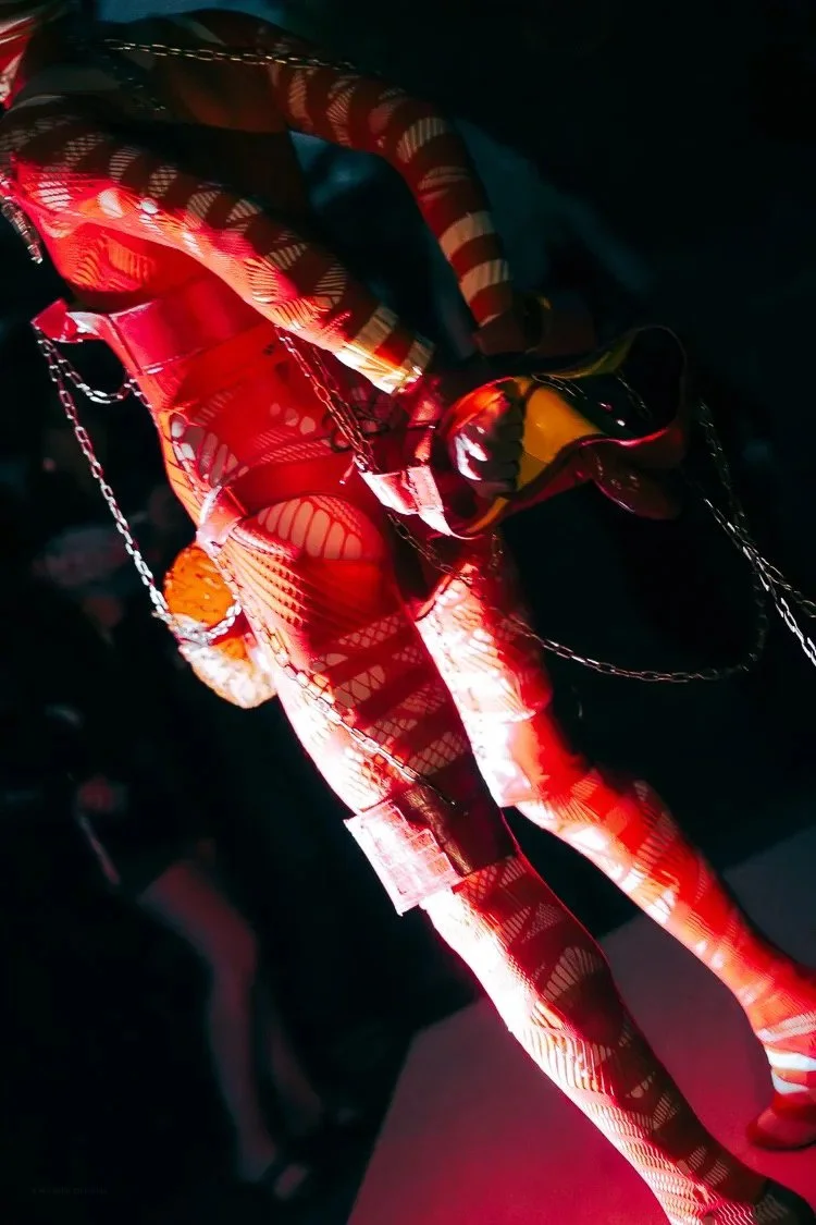 Performer dressed in a vibrant red and gold patterned costume with chains, holding a black and yellow mask, illuminated by stage lighting.