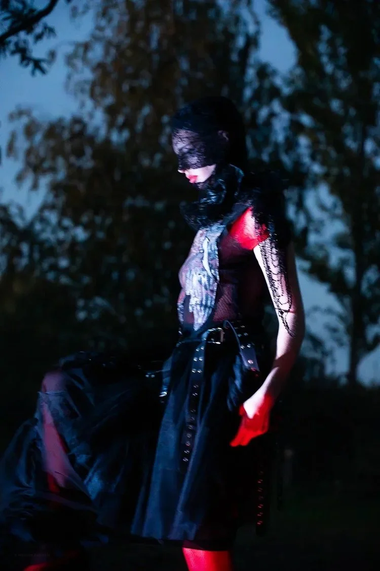 A woman dressed in dark clothing with a lace mask covering her eyes, standing outdoors at dusk or night with blurred trees in the background, illuminated by a red-tinted light.