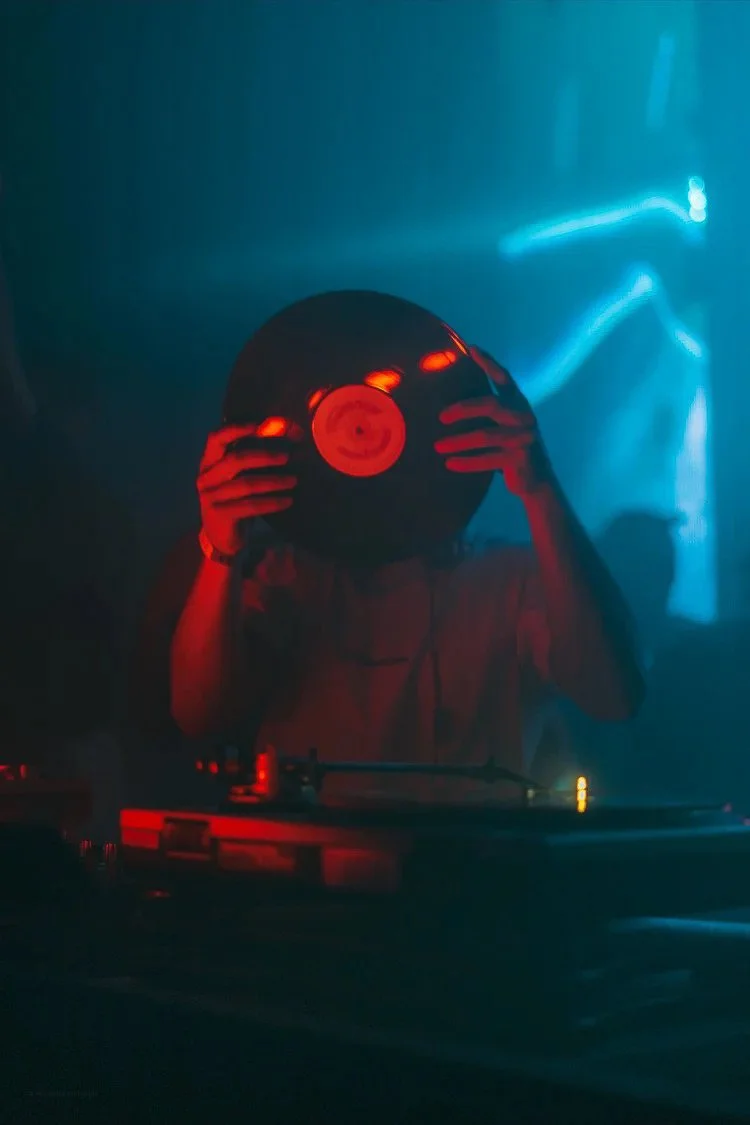 Person DJing at a nightclub, holding a vinyl record in front of their face, with blue and red lighting.
