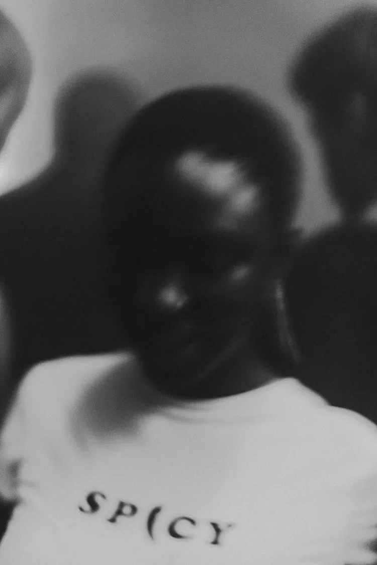 A black and white photo of a smiling person wearing glasses and a T-shirt with the word 'SPICY' printed on it, holding up two fingers.