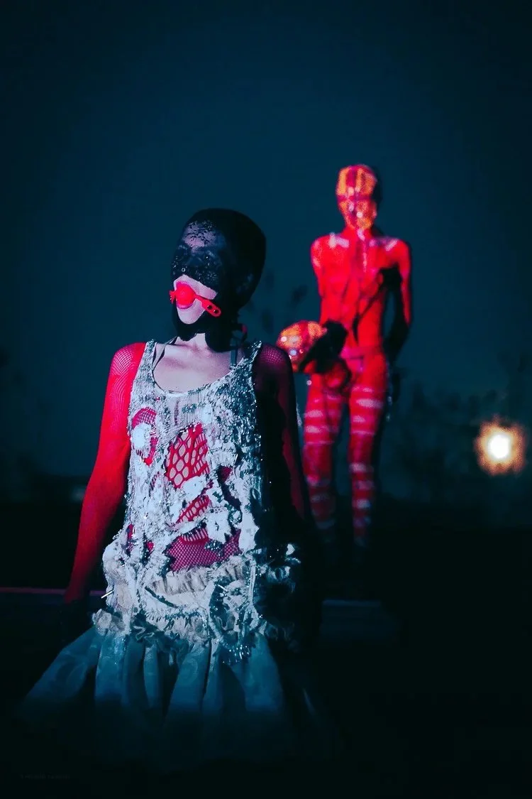 Two mannequin-like figures with mannequin-like heads, one in the foreground and one in the background, wearing red and patterned clothing, illuminated in dark lighting.