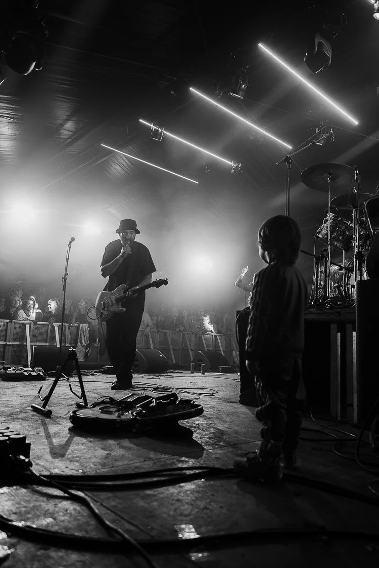 A musician with a guitar performing on stage in a black and white photo, with a drummer and audience in the background, stage lights overhead, and various equipment on the floor.