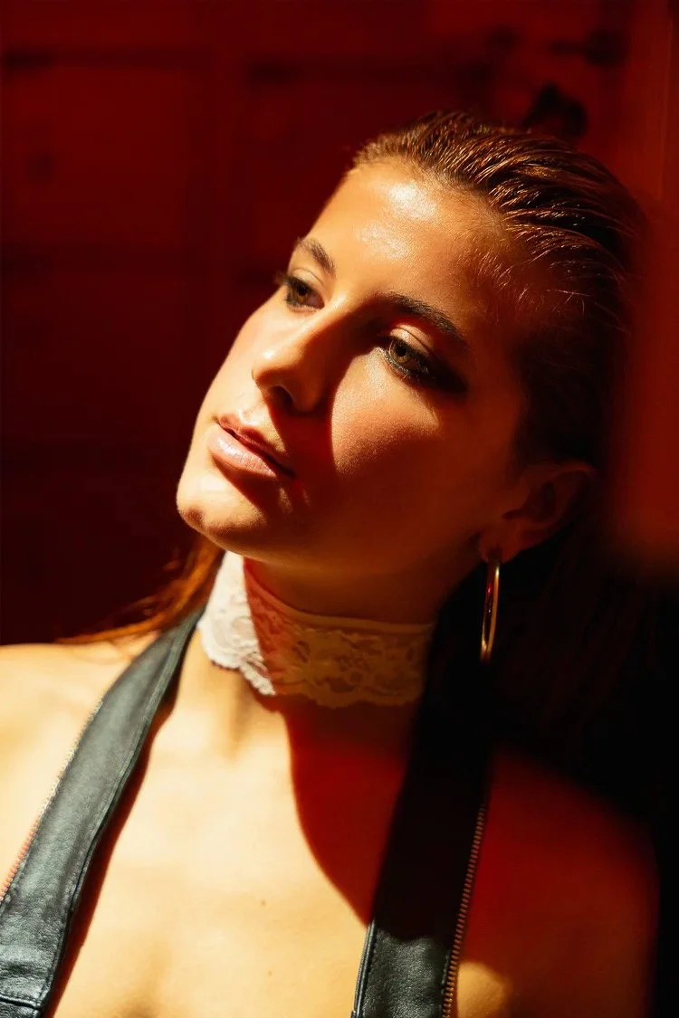 A young woman with long hair and hoop earrings looking to the side, illuminated by warm lighting against a red background.