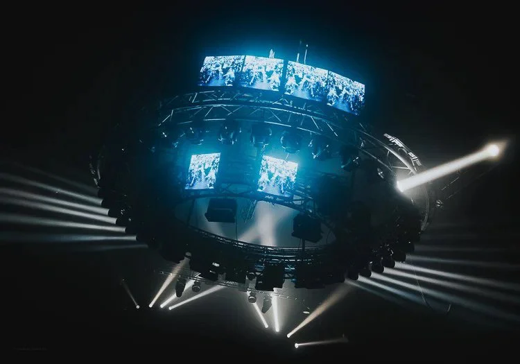 A circular stage or lighting rig with multiple screens displaying visuals, surrounded by bright spotlights and beam lights in a dark concert or event setting.
