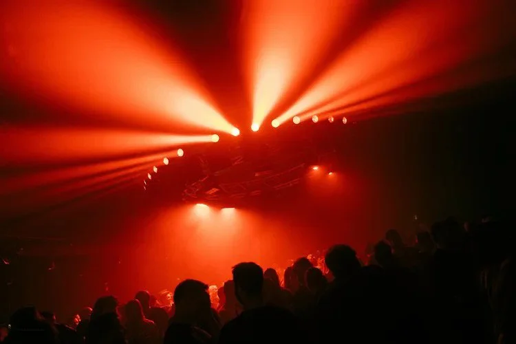 A concert or nightclub scene with silhouetted people facing a stage illuminated by intense red and orange lights.