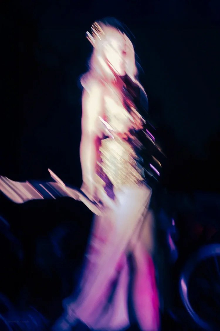 Blurry image of a woman standing on stage with a microphone, wearing a shiny dress and a crown, with stage lights creating a colorful effect.