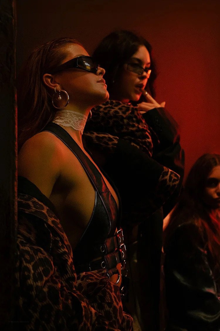 Three women standing against a red wall, wearing dark clothing and sunglasses, with the woman in the foreground wearing a lace choker and hoop earrings.