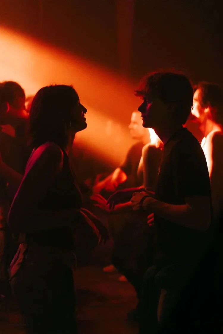 Silhouettes of young people dancing and socializing at a dimly lit party or nightclub, illuminated by warm red and orange lighting.