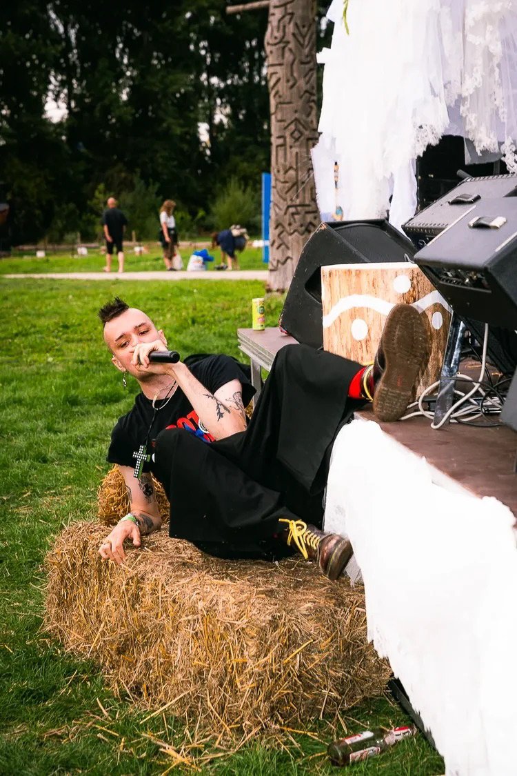 A person with a mohawk hairstyle, tattoos, and a cross necklace is lounging on a hay bale at an outdoor event, holding a microphone near their mouth. They are dressed in black pants and a t-shirt, with yellow shoelaces, and appear relaxed. In the bac
