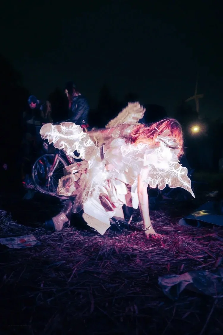A person dressed in a flowing, luminous costume performing or posing outdoors at night, with a bicycle and other people visible in the background.