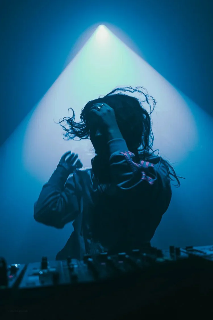 A woman with curly hair, wearing a jacket, is DJing in a dark, blue-lit setting with a spotlight above her.