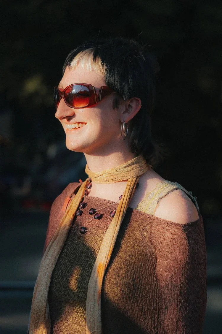 A woman with short, dark hair with light streaks, wearing large sunglasses, hoop earrings, a multicolored top, and a choker around her neck. She is smiling and standing outdoors in sunlight.