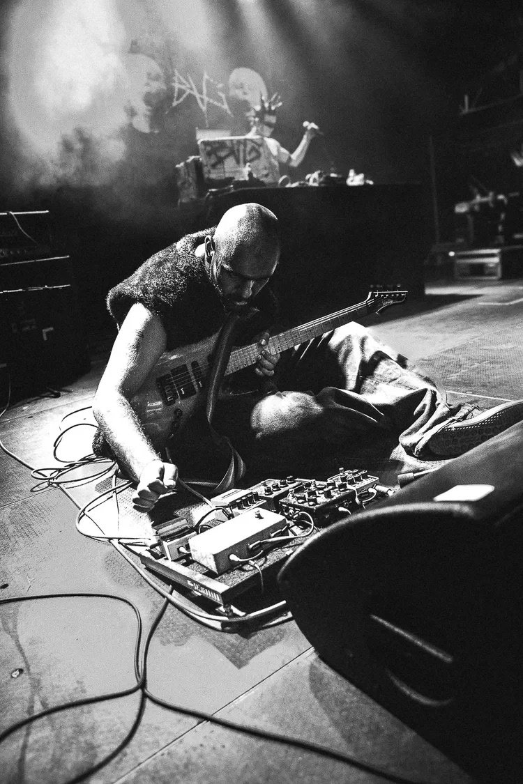A musician kneeling on the stage floor with a guitar and music equipment, while another person performs DJing in the background during a live music event.