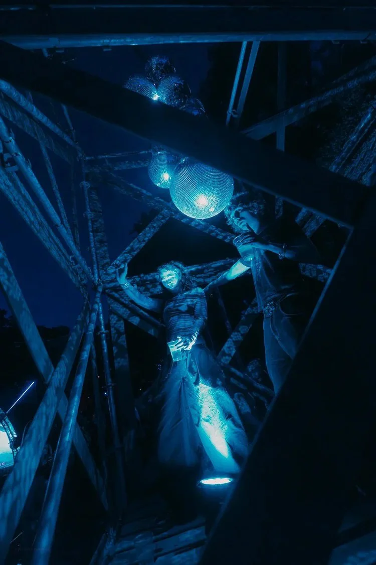 Two people wearing reflective masks on a metal structure with disco balls and blue lighting.