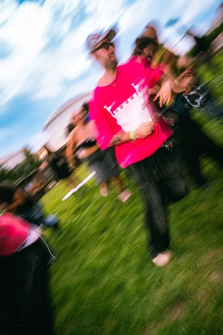 A man with a beard, wearing sunglasses and a pink T-shirt, walking outdoors on grass during daytime, with several other people blurred in motion around him.