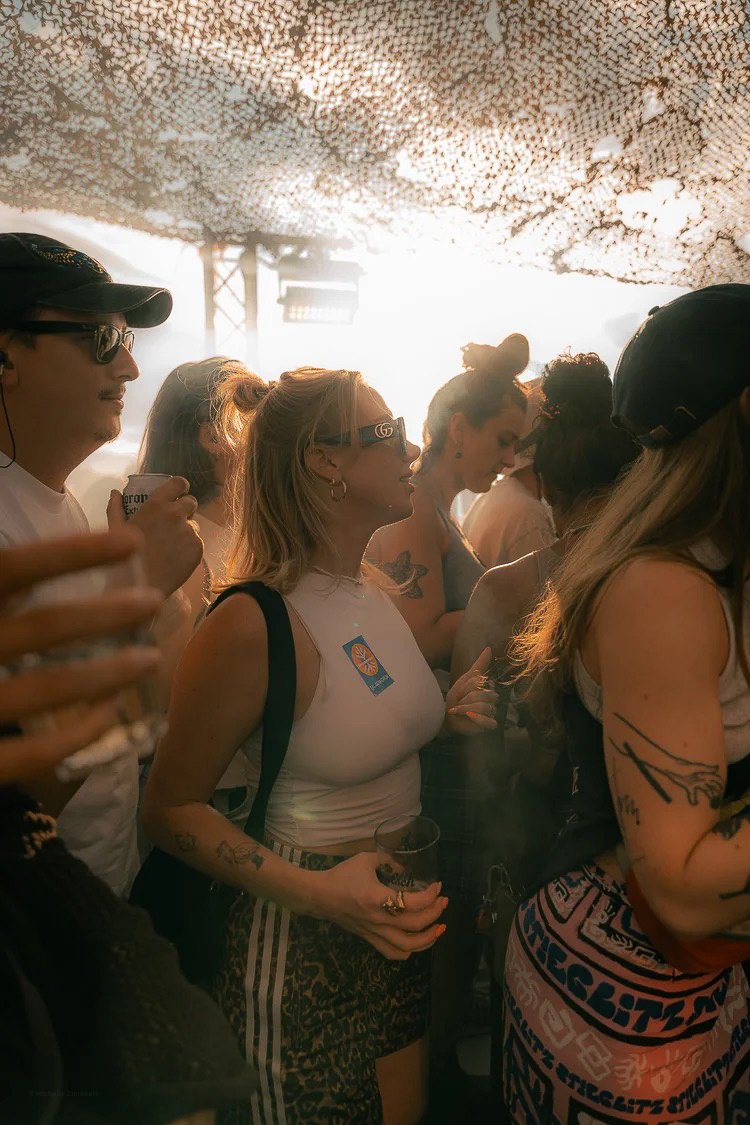 A group of young adults at an outdoor event, standing under a canopy, with warm sunlight in the background. They are wearing casual clothing, some with sunglasses and tattoos, holding drinks, and appear to be enjoying a social gathering.