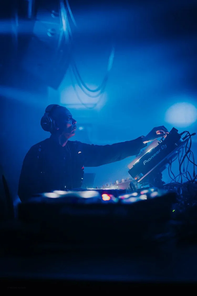 DJ performing at a nightclub or music event with blue lighting, mixing music on a Pioneer DJ controller.