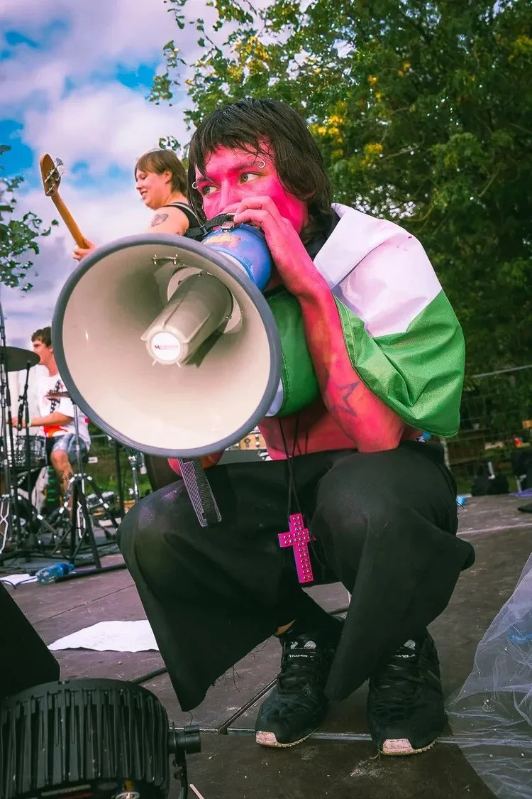 Person with pink face paint and green, white, and black clothing speaking into a megaphone on a stage outdoors, with other people and musical equipment in the background.