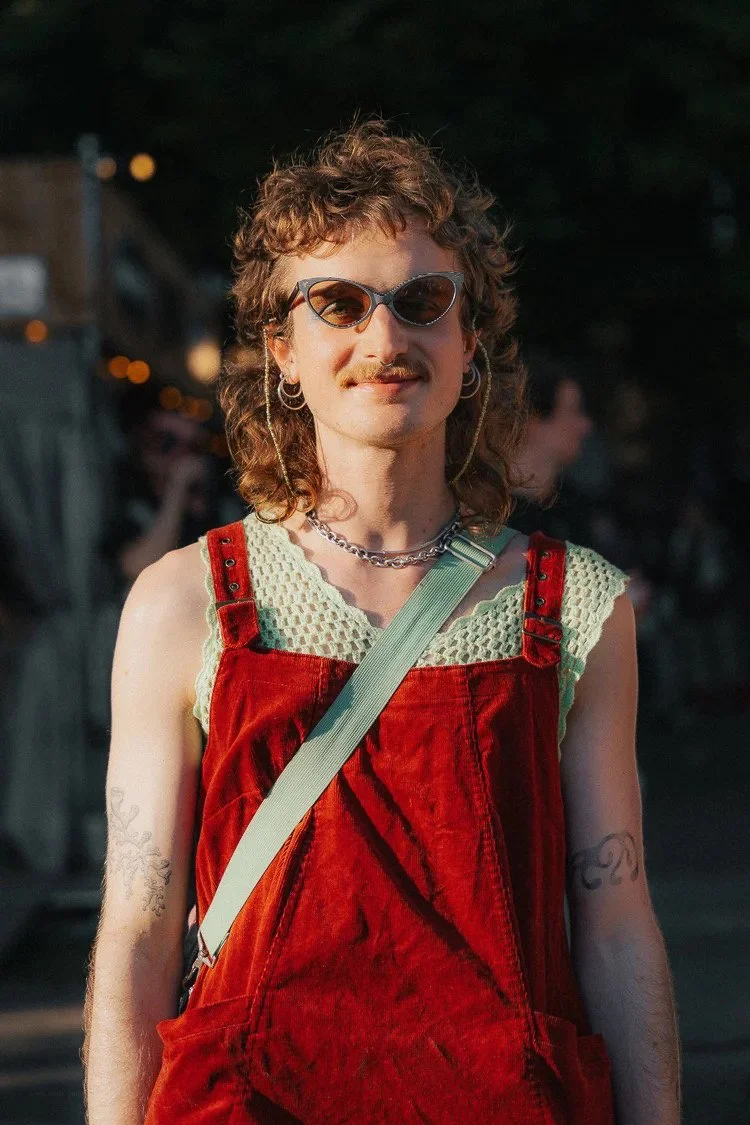 A person with curly hair, sunglasses, and hoop earrings is standing outdoors, wearing a red jumper over a crochet top and has tattoos on their arms.