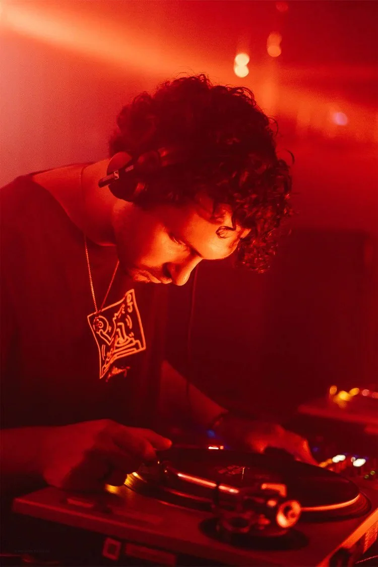 DJ with curly hair wearing headphones and a black shirt, playing music on a turntable under red lighting.