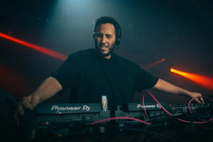 A male DJ wearing headphones operating DJ equipment during a live performance with red and orange lighting effects.