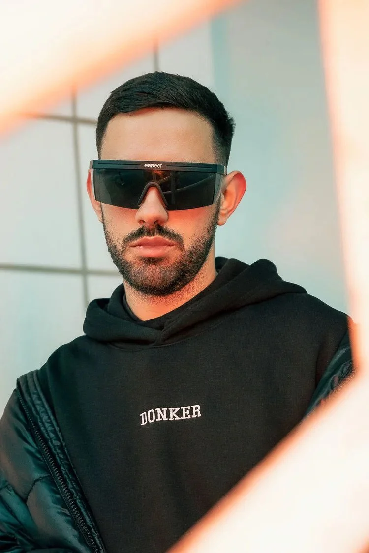 A young man with short dark hair and a beard, wearing large black sunglasses and a black hoodie with the word "DONKER" on it, looks into a mirror with a reflection of a bright light on the top left corner.