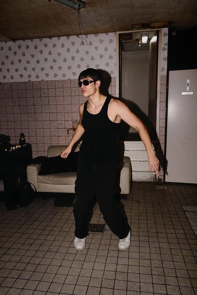 Person in black tank top, black pants, white sneakers, and sunglasses dancing in a room with tiled floors and patterned walls.