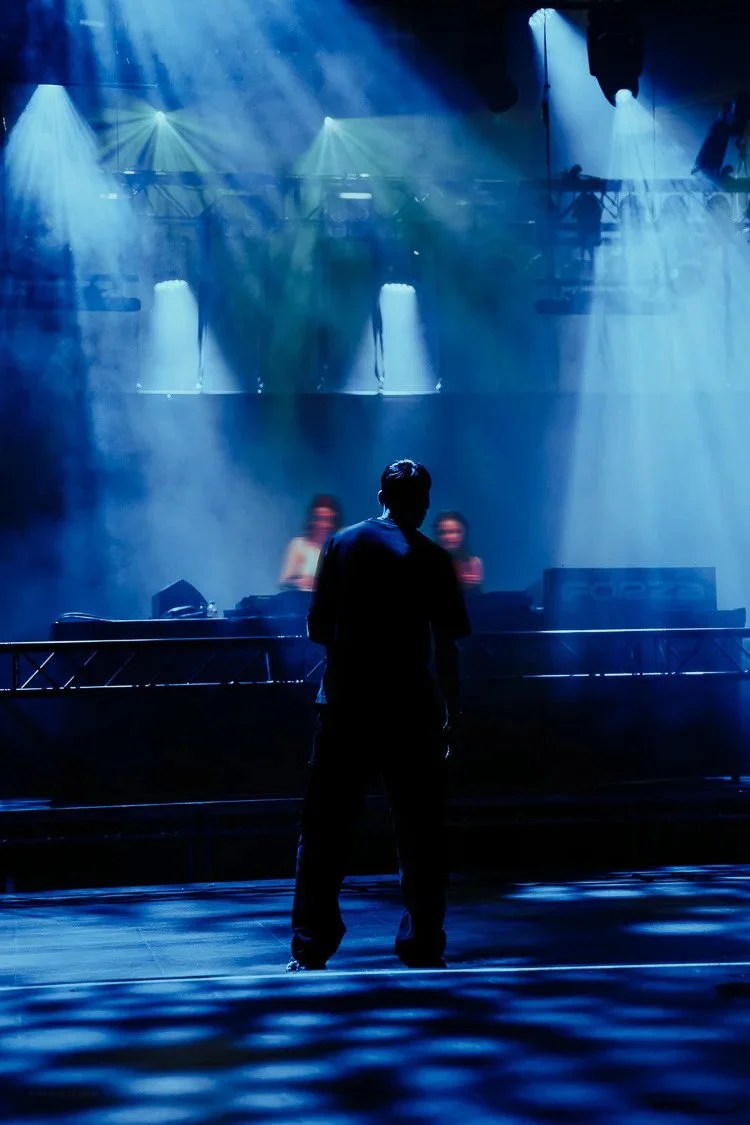 A person standing in front of a stage with two women DJing behind a DJ booth, illuminated by blue stage lights.