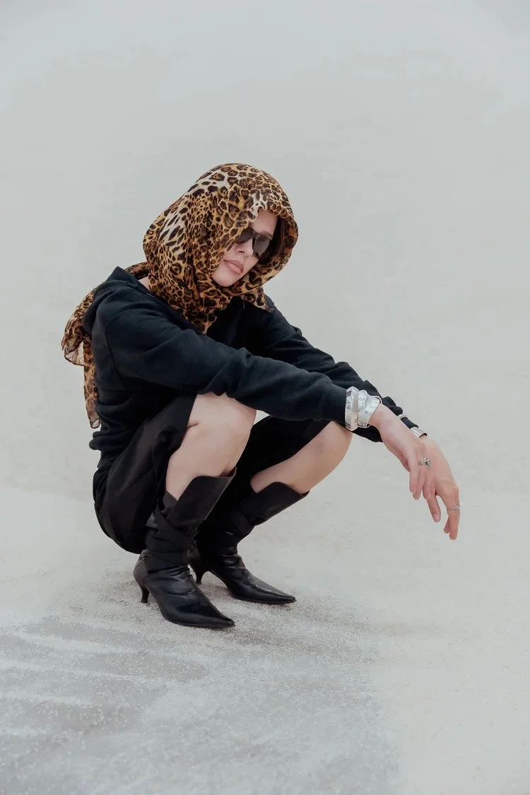 Woman wearing leopard print headscarf, sunglasses, black outfit, and high-heeled boots squatting on sandy ground.