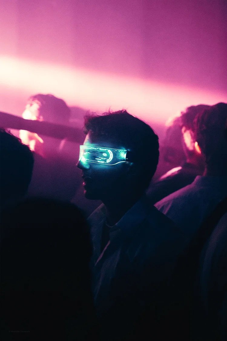 Person wearing glowing virtual reality glasses at a crowded indoor event with pink and purple lighting.