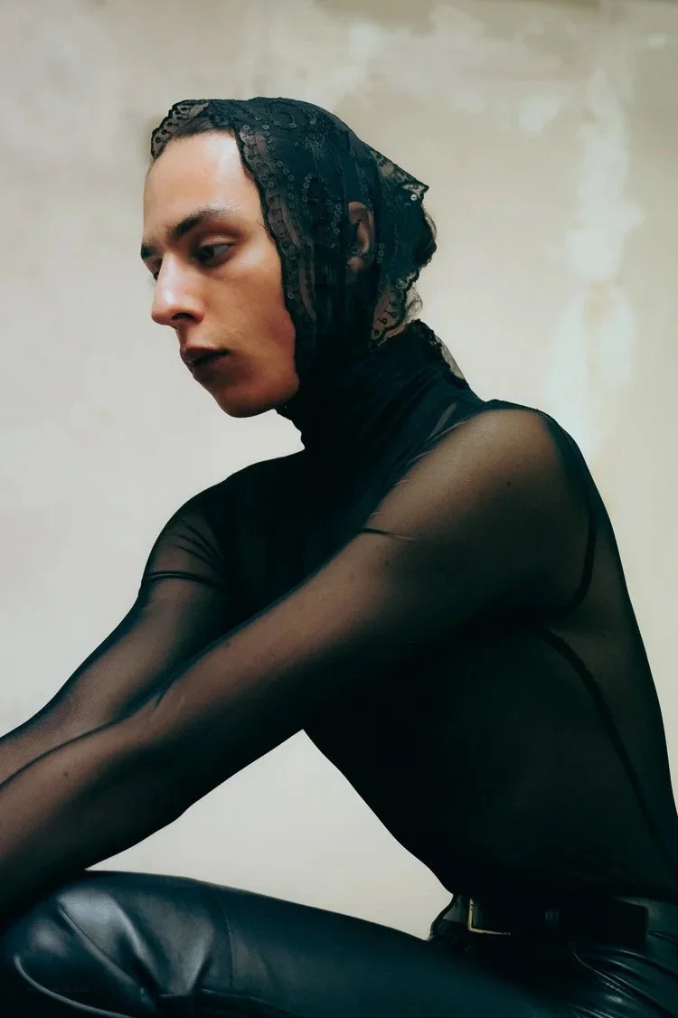 Person wearing a black lace head covering and a sheer black top, sitting with arms resting on knees and looking down.