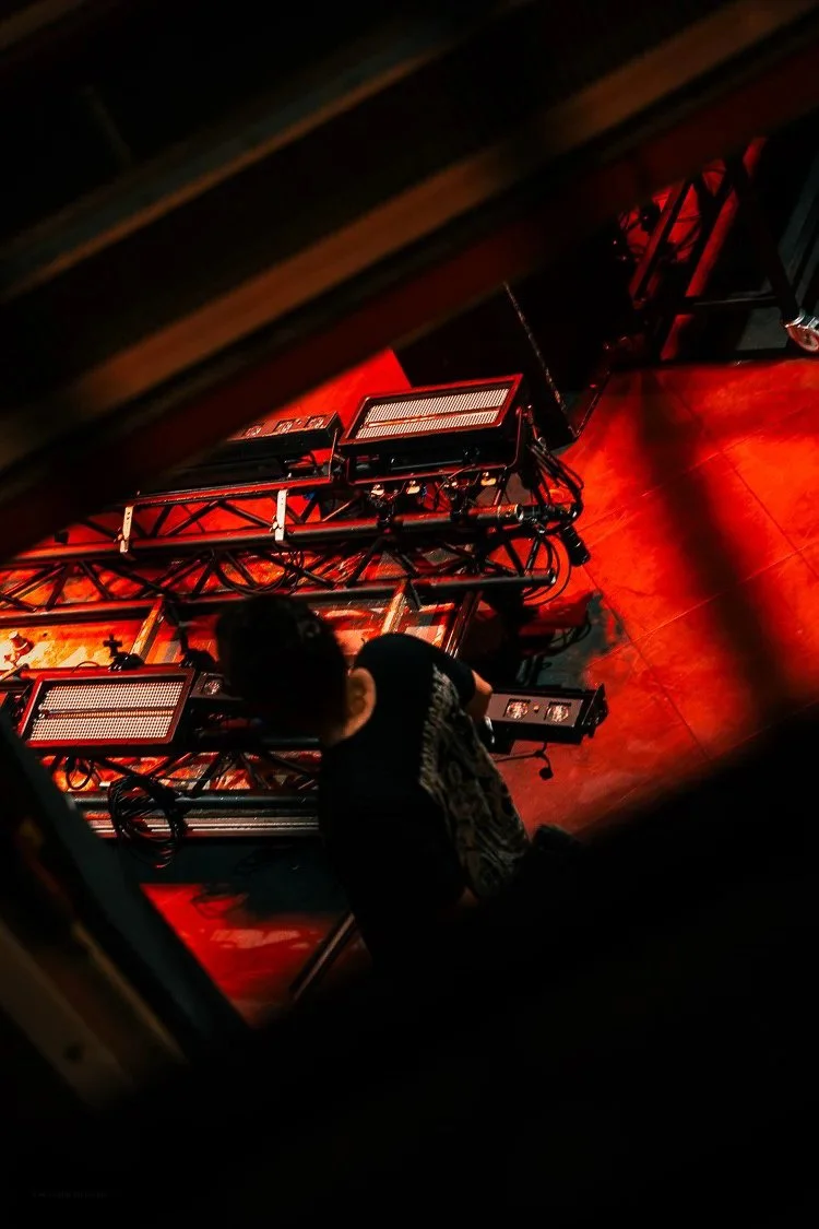 A person kneeling on a stage with lighting equipment and scaffolding, viewed through a gap in a wooden structure, with stage lights casting a red glow.