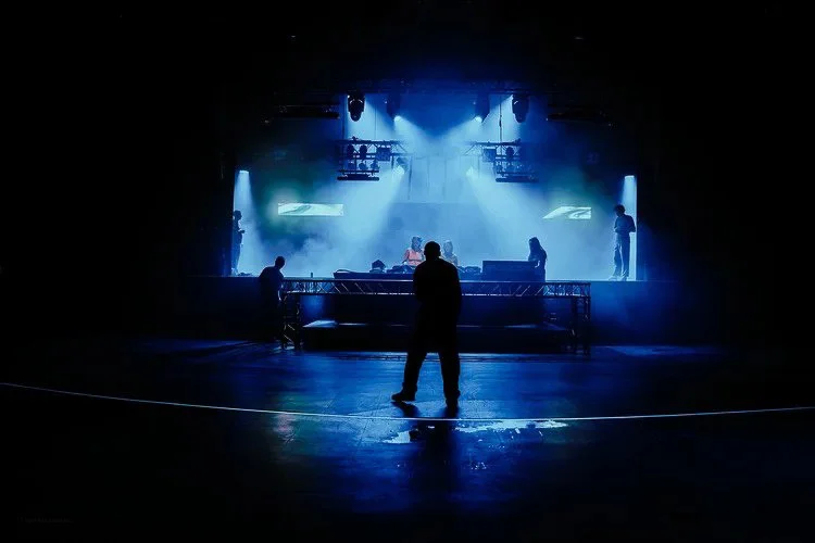 People performing on a stage with bright blue spotlights, seen from the audience in a dark venue.