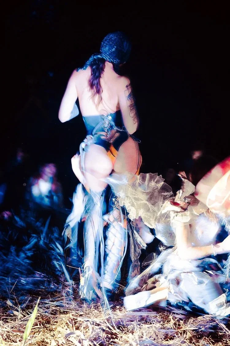 A woman dressed in dark clothing and fishnet stockings, wearing a hood, walking away at night in an outdoor setting with dark sky and grass.