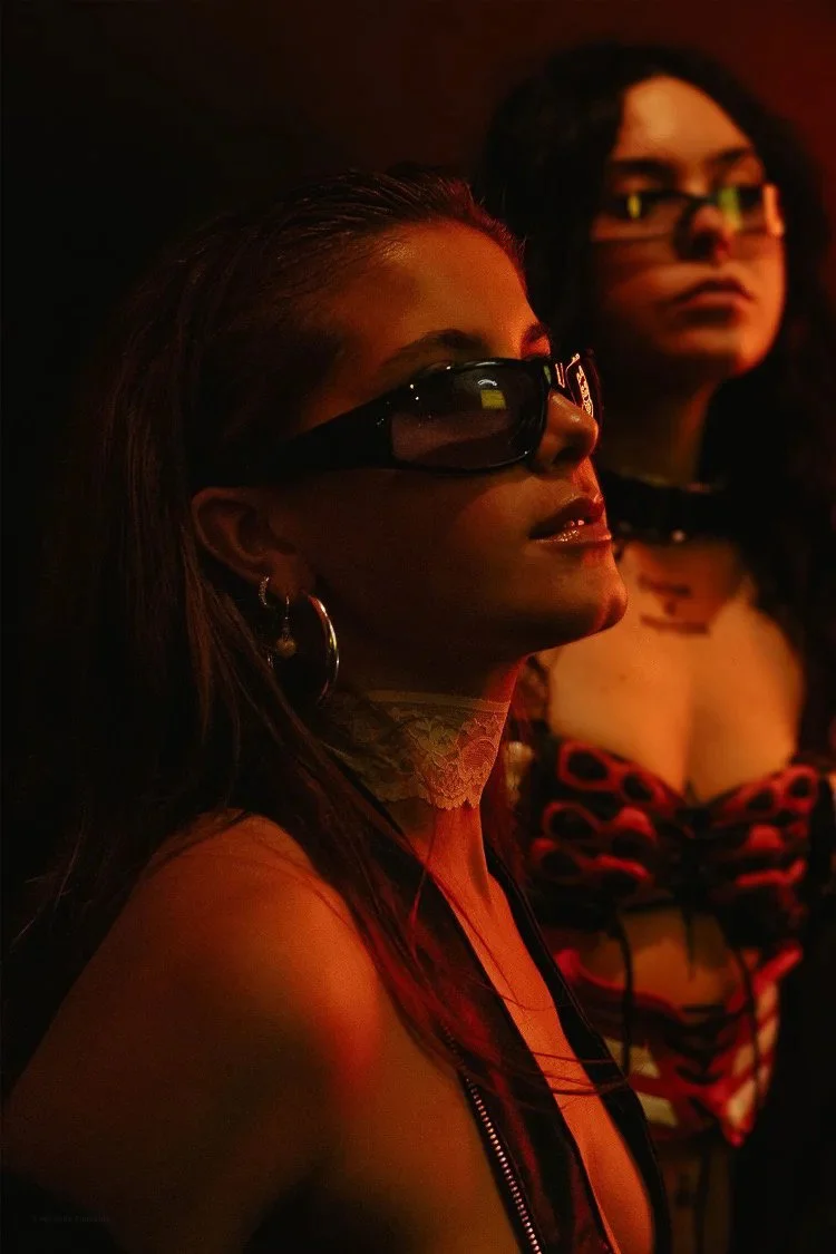 Two women wearing sunglasses, one with earrings and necklace, in a dimly lit setting with warm red and orange lighting.
