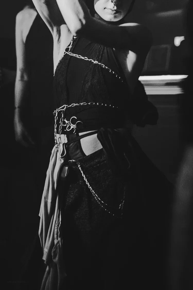 A black and white photo of a woman wearing a black top with chain details, with one arm raised, in a dark setting with faint background lights.