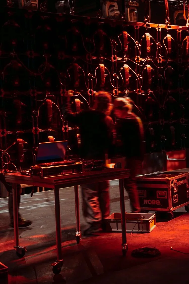 Two individuals working on sound equipment in a dimly lit stage production or concert setting, with a wall of electronic gear behind them.