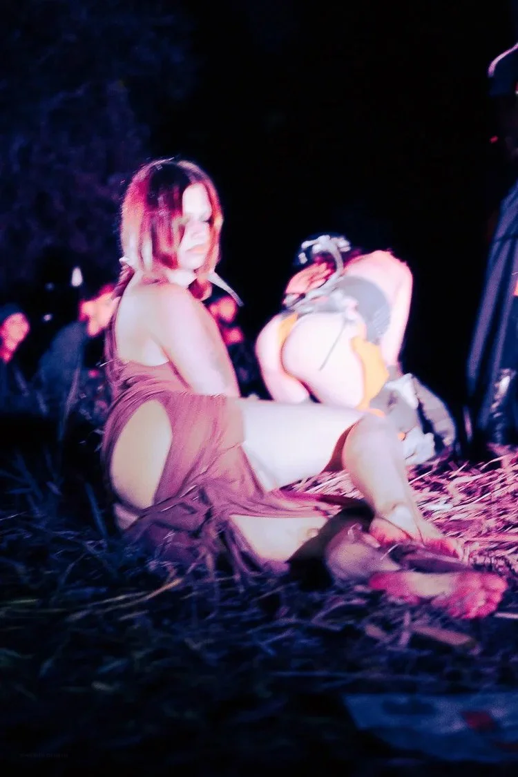 A woman sitting outdoors at night, dressed in a revealing outfit, with people and trees in the background.