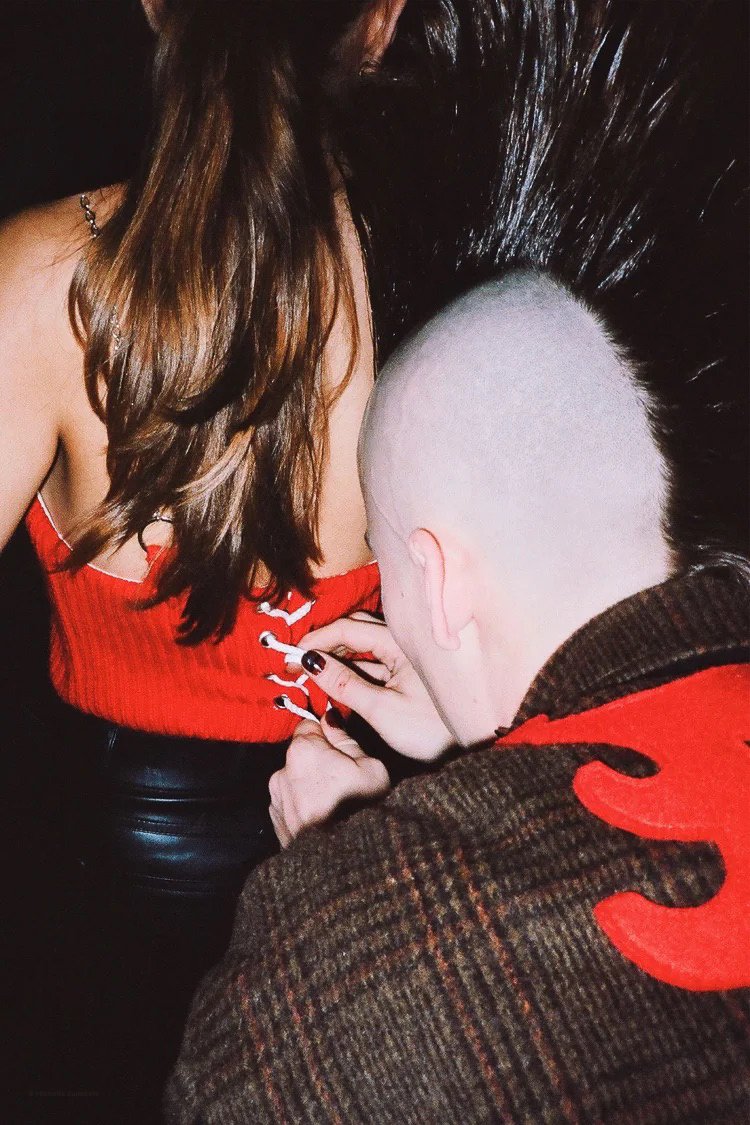 A man with a shaved head and fair skin is sewing eyelets onto the red top of a woman with long, brown hair and darker skin. The woman is wearing a red tank top with black pants, and the man is wearing a brown plaid jacket with a red flame design on t