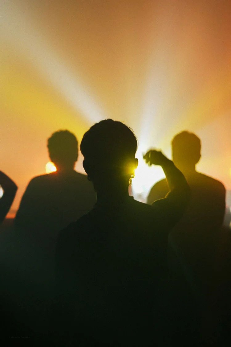 Silhouettes of three people against a vibrant background with bright lights and beams, possibly at a concert or outdoor event during sunset or sunrise.